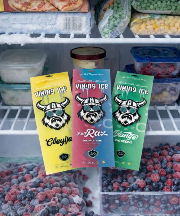 Multiple flavors of Viking Ice 100mg THC freezer pops laying flat on a freezer shelf next to frozen fruit and vegetables.